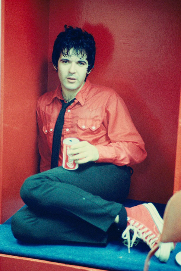 A color photo of a Clem Burkle from Blondie sitting in a red chair, wearing a red shirt and green pants, holding a drink.