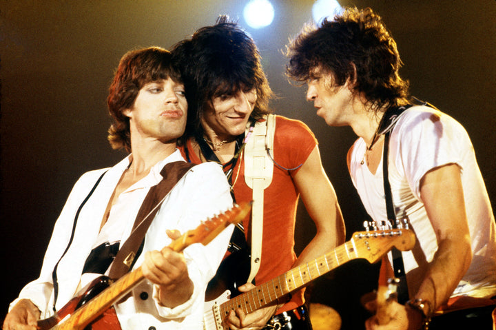 A classic photograph of The Rolling Stones featuring members Mick Jagger, Keith Richards, and Ron Wood, with a fourth member holding a guitar. The photo captures a moment from a concert with a bright light in the background.